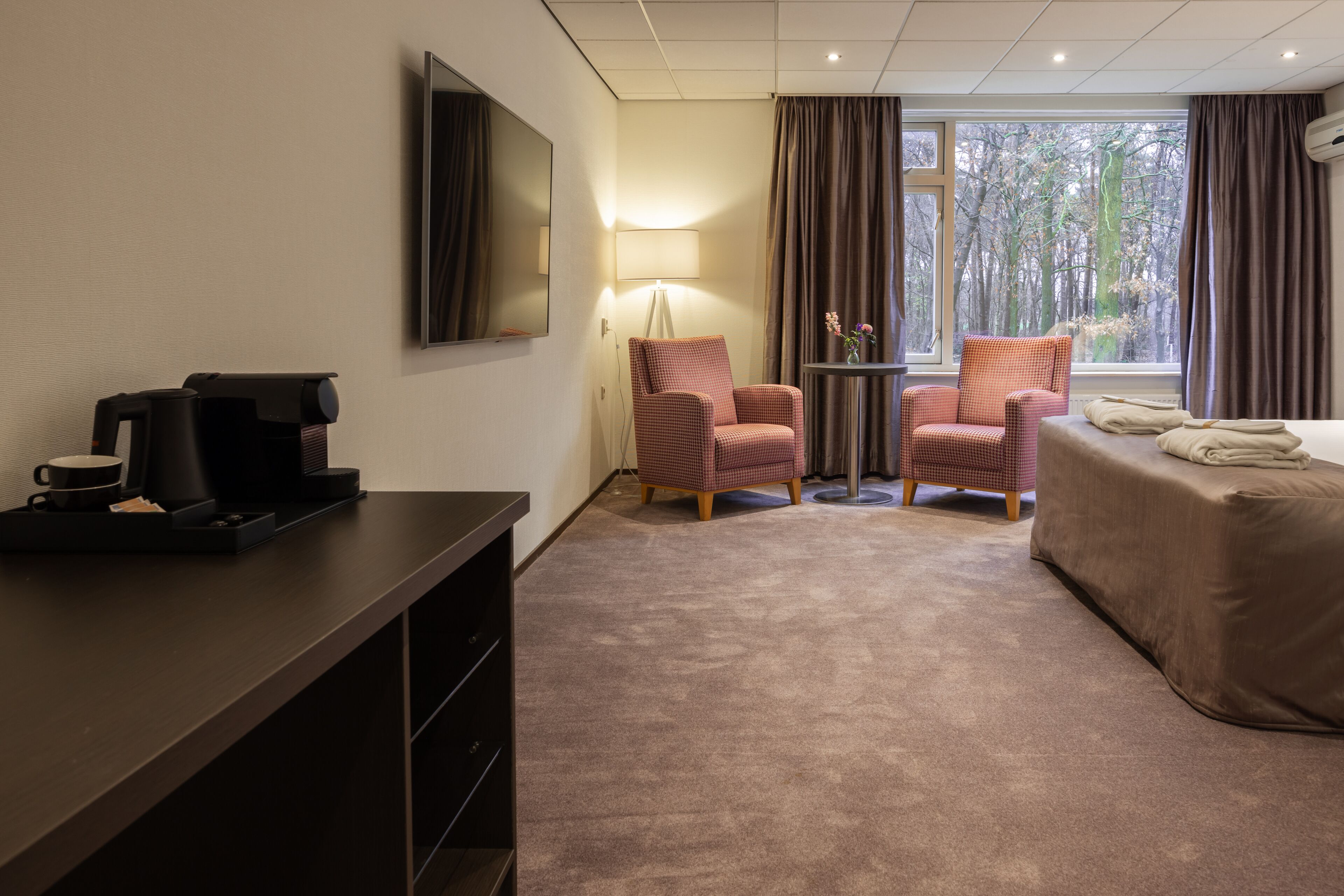 A hotel room with a bed, two armchairs, a coffee maker, a lamp, and a window with curtains.