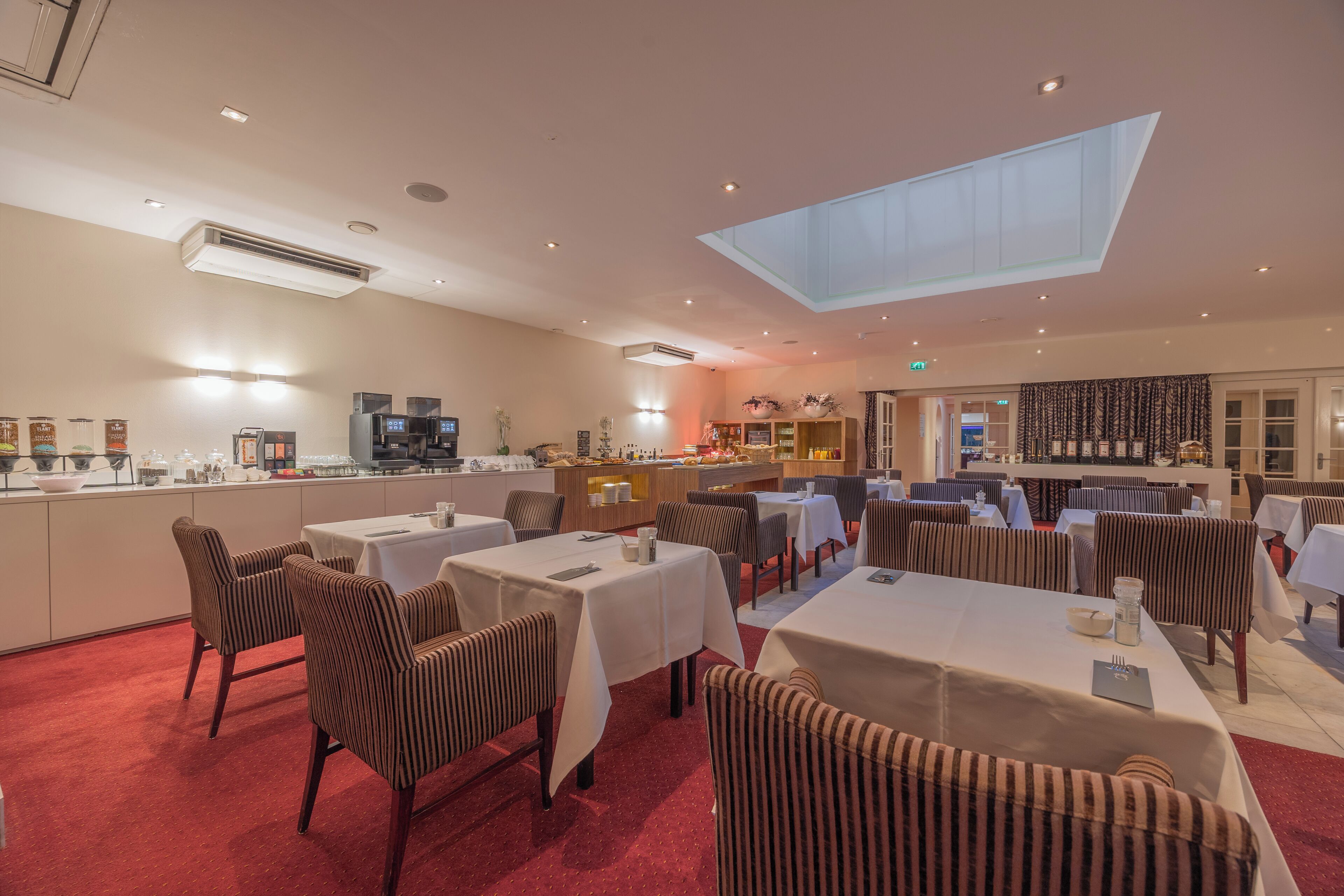 Un restaurante con mesas, sillas, una zona de cocina y un tragaluz.