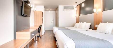 Ein modernes Hotelzimmer mit einem Holztisch, zwei Betten mit weißer Bettwäsche, einem an der Wand montierten Fernseher und einer kleinen Küchenzeile.