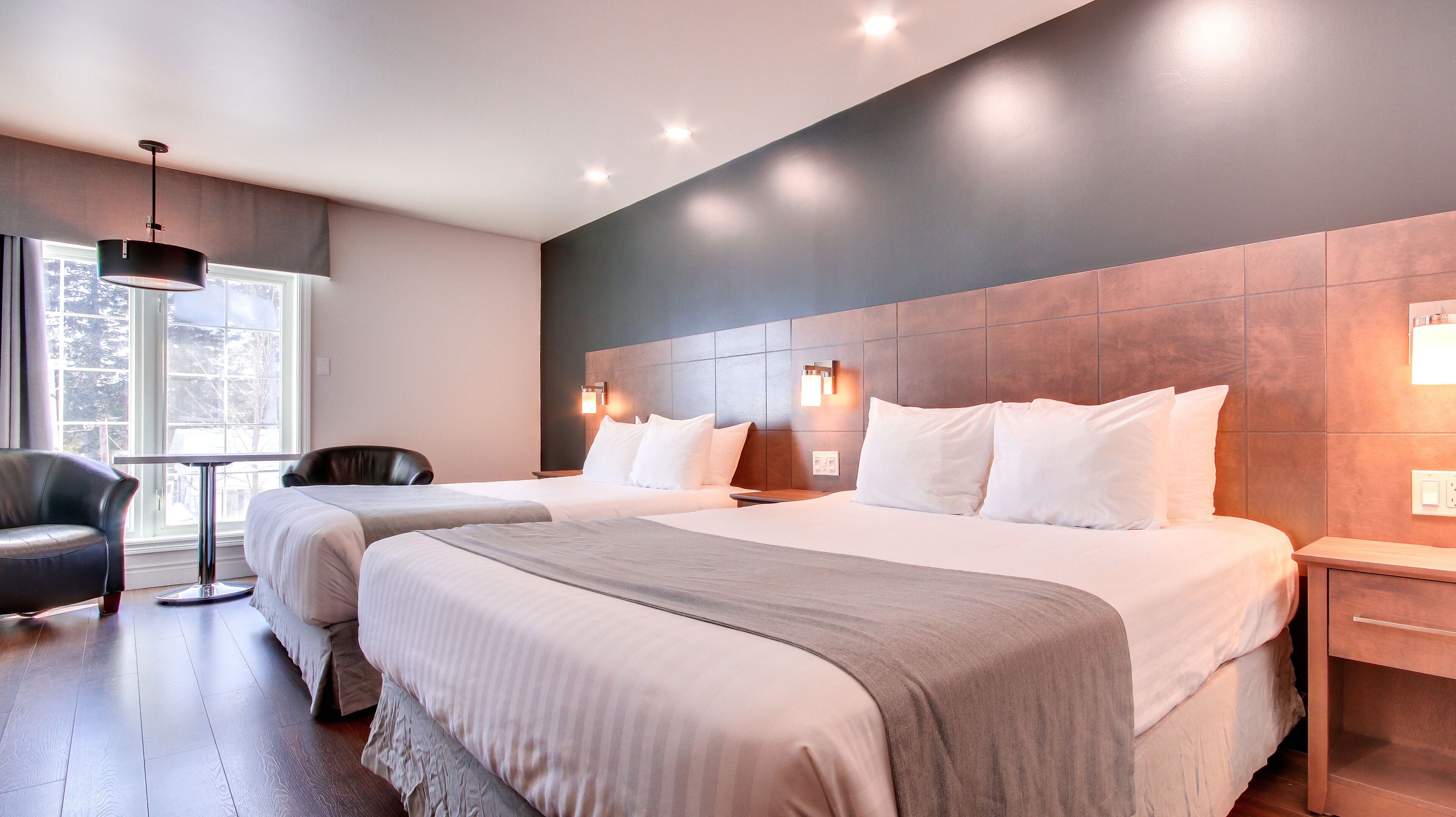 A modern hotel room with two beds, a large window, a leather chair, and a wooden nightstand.