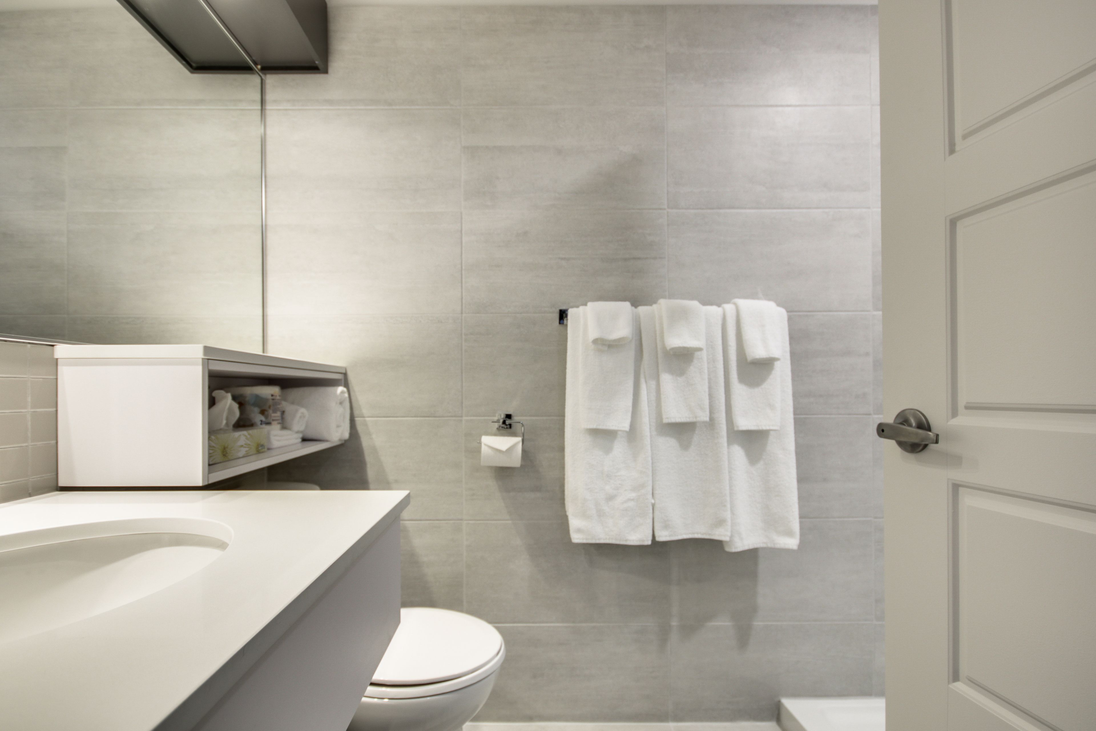 Un baño moderno con lavamanos, inodoro y zona de ducha blancos. Toallas blancas cuelgan de un perchero.