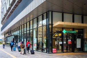 Une entrée d’hôtel moderne avec des portes en verre, une enseigne bien visible indiquant « HANSHIN », et des personnes avec des bagages à l’extérieur.
