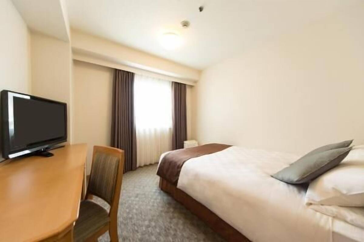 A hotel room with a bed, a desk with a television, a chair, and a window with curtains.