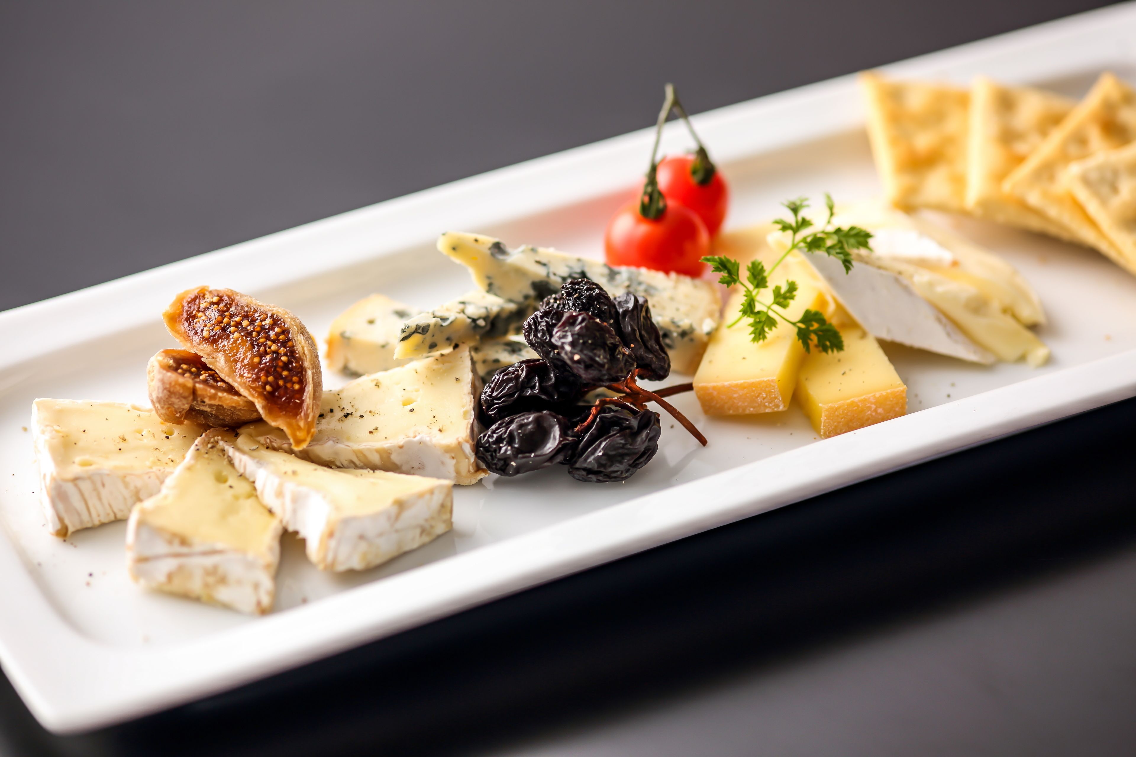 Une assiette rectangulaire blanche sur laquelle on trouve divers fromages, des fruits secs et une tomate cerise.