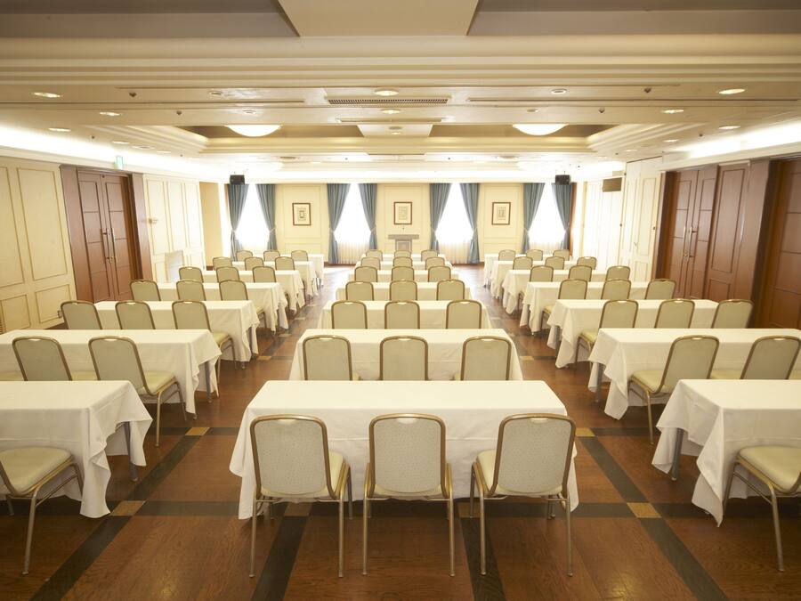 A conference room with rows of tables and chairs, large windows with curtains, and a podium at the front.
