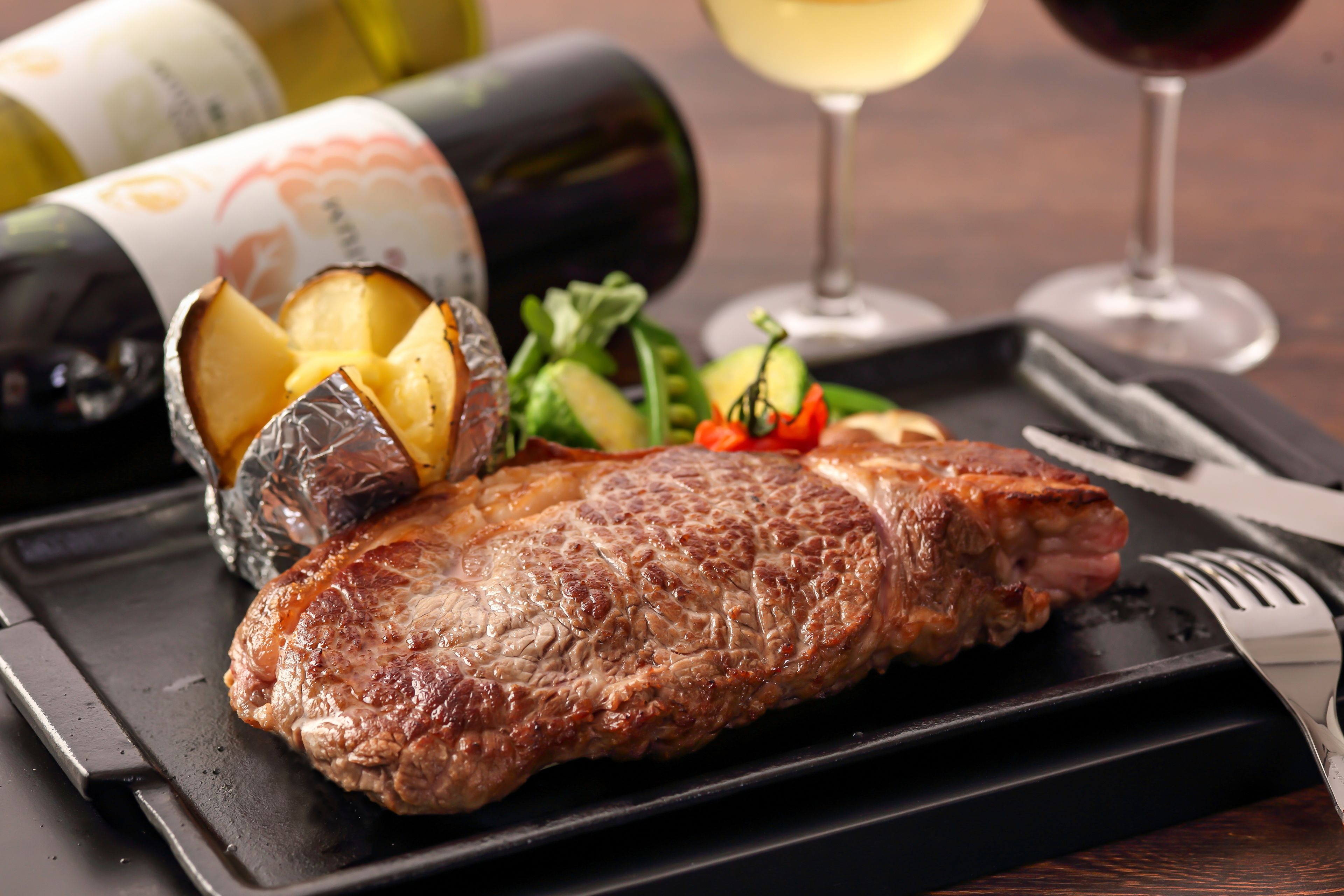 Un plato negro con carne, papas asadas y vegetales, acompañado de botellas y copas de vino.