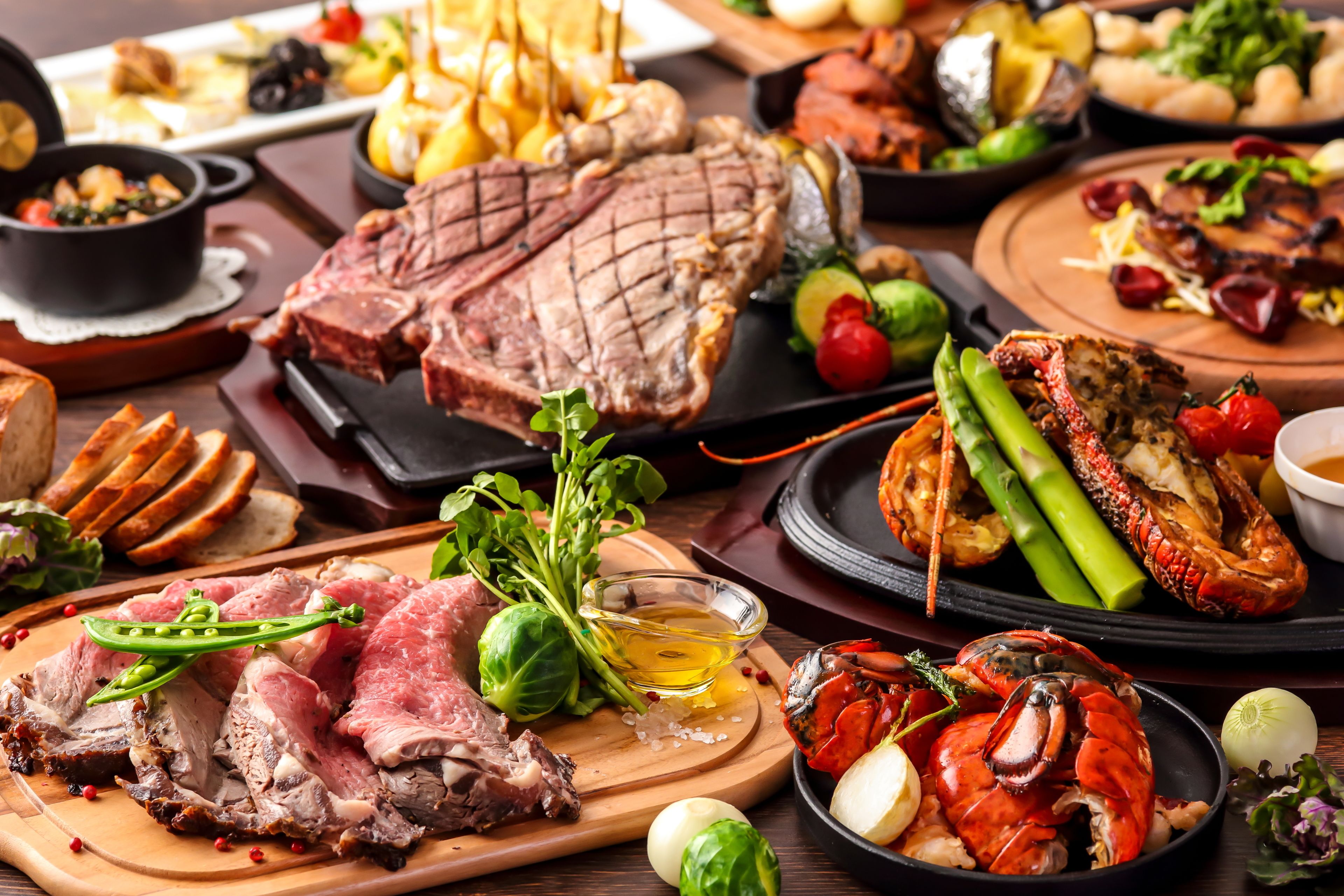 A variety of meats including steak and lobster, accompanied by vegetables and bread on a wooden board.