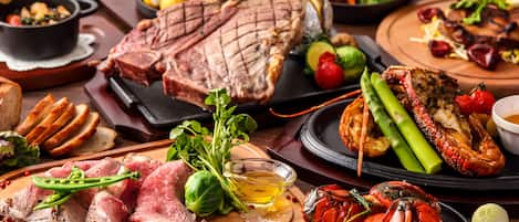 A variety of meats including steak and lobster, accompanied by vegetables and bread on a wooden board.