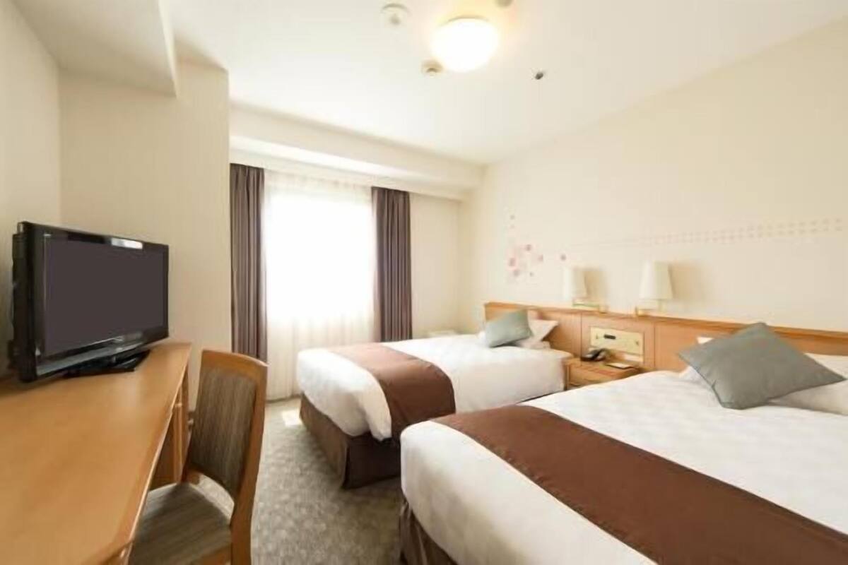 A hotel room with two beds, a desk, a television, and a window with curtains.