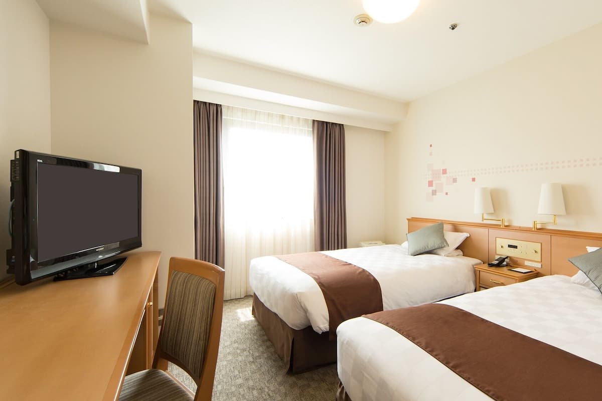A hotel room with two beds, a desk with a television, a chair, and a nightstand with lamps.