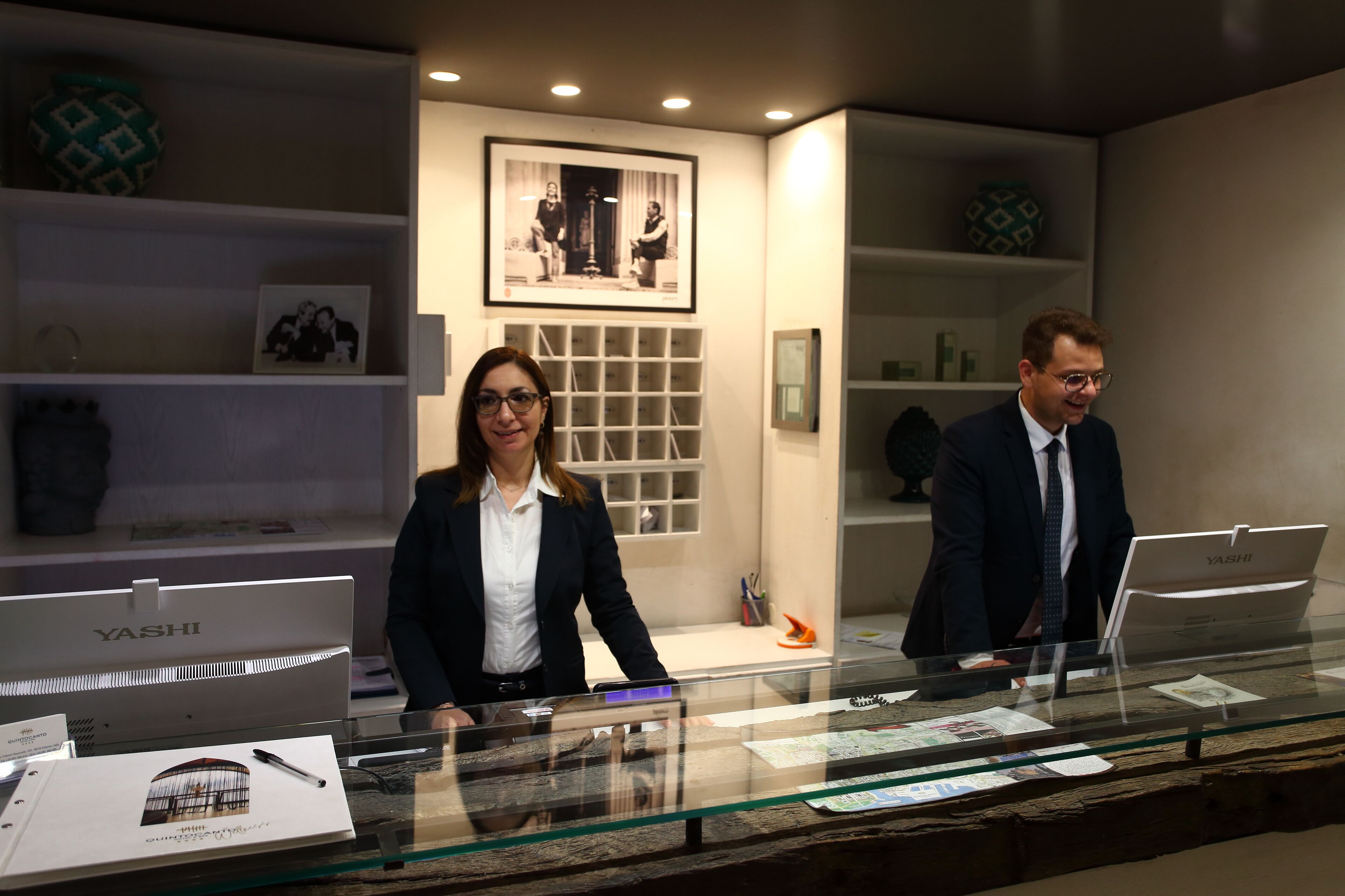 A reception desk with a glass partition, a framed photograph on the wall, and a YASHI laptop on the counter.