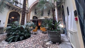 Un patio con suelo de piedra, sillas amarillas, plantas en macetas y la entrada a un edificio.