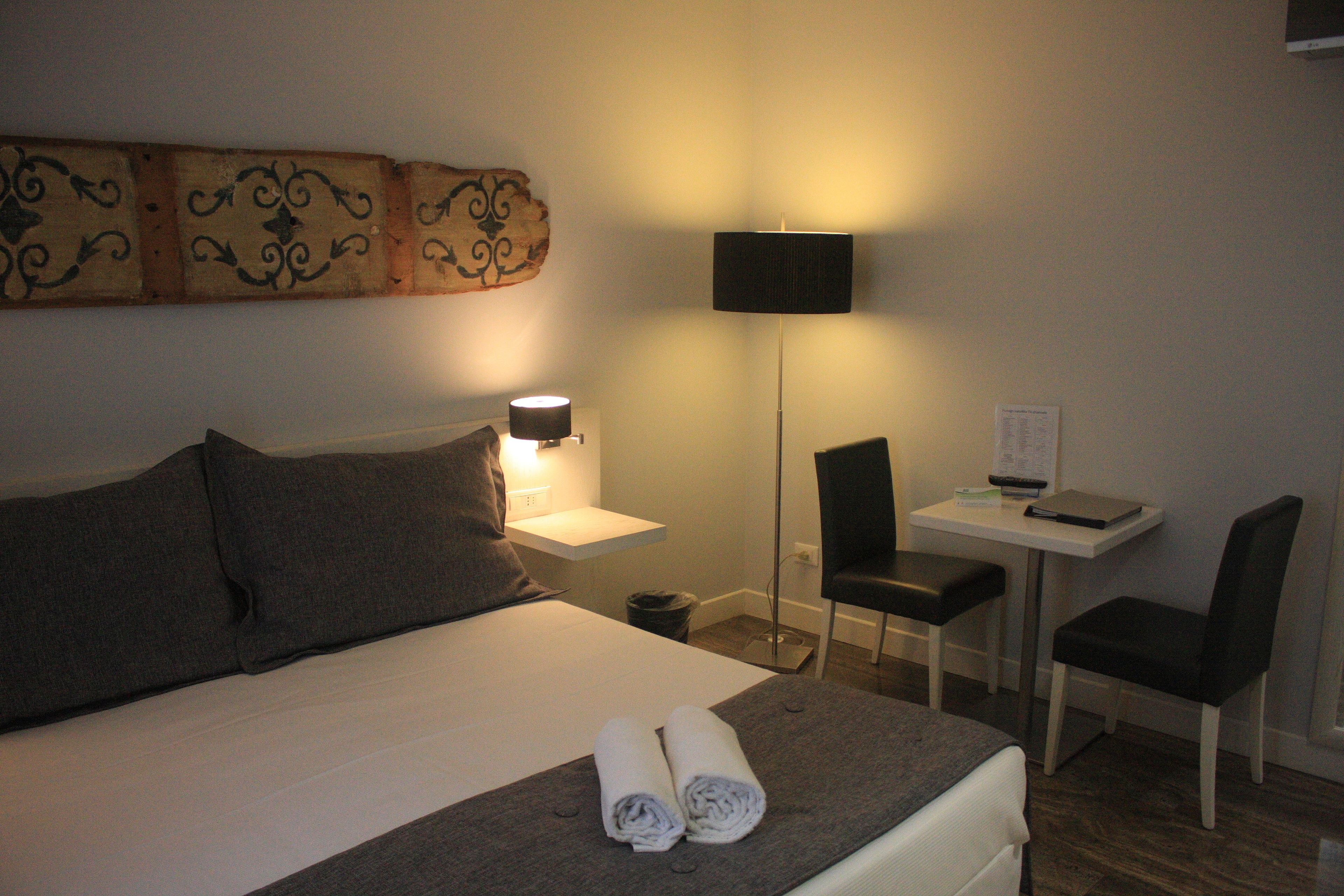 A hotel room with a bed, two pillows, a bedside table with a lamp, a desk with a chair, and a wall-mounted artwork.