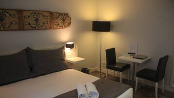 A hotel room with a bed, two pillows, a bedside table with a lamp, a desk with a chair, and a wall-mounted artwork.