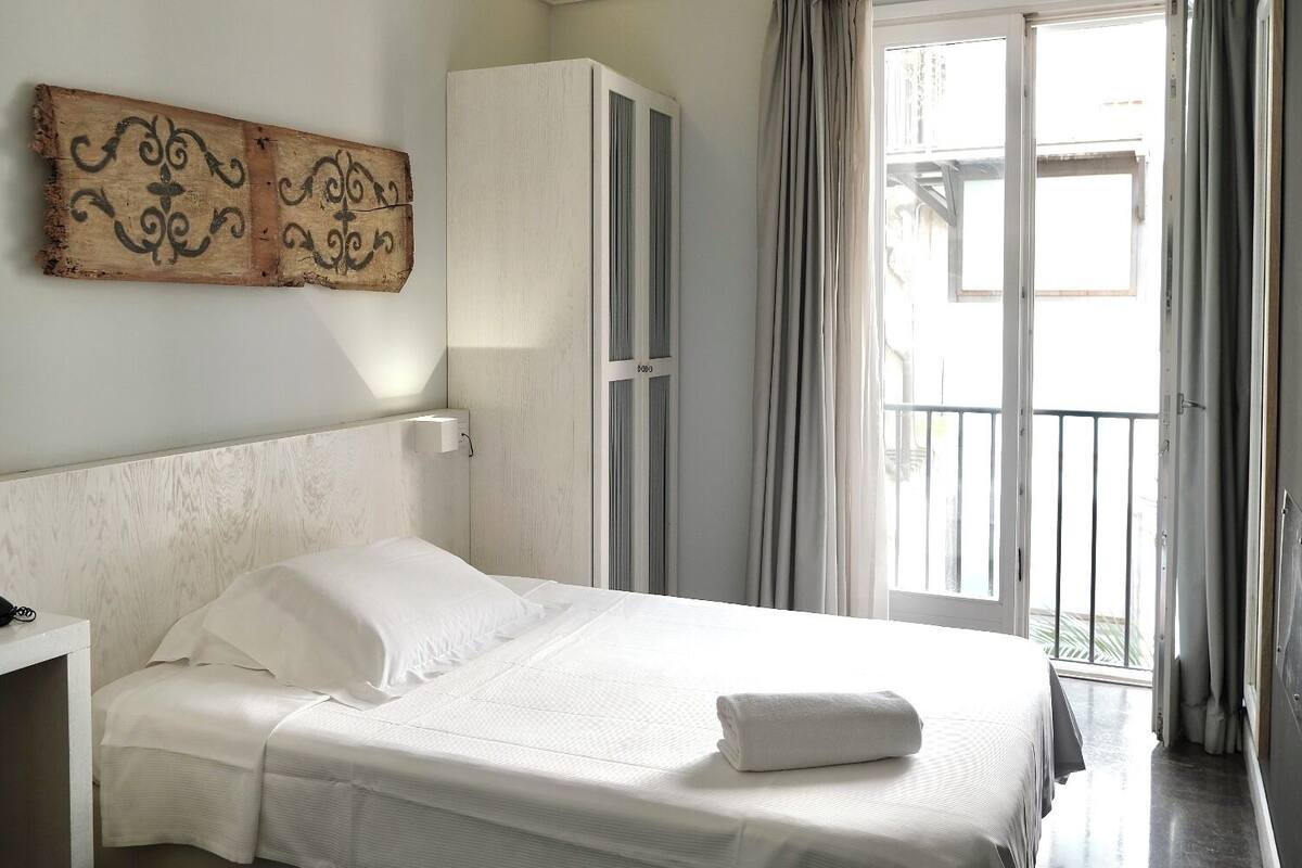 Une chambre d’hôtel comprenant un lit, un bureau avec un téléphone, une armoire et un balcon avec des rideaux.