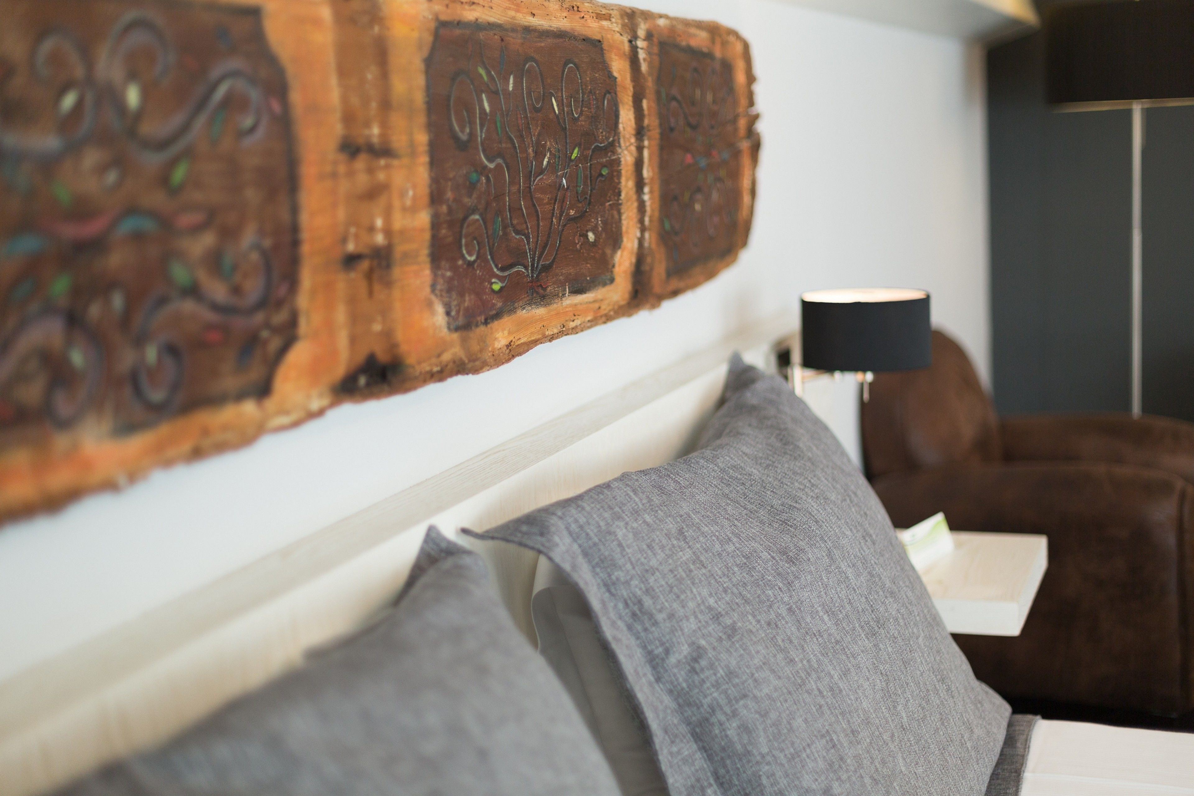 Une chambre avec une tête de lit en bois, des coussins gris, un fauteuil en cuir marron et un lampadaire.