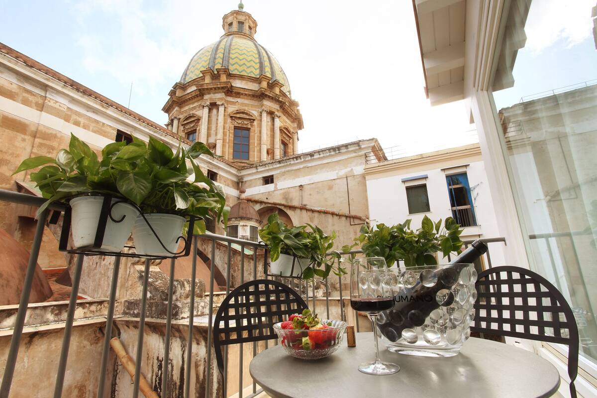 Un balcon agrémenté de plantes en pot, une table avec un saladier de fruits et un verre de vin, et une vue sur un bâtiment historique surmonté d’un dôme.