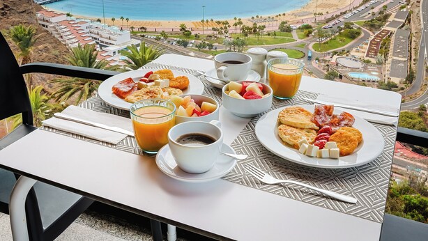 A table with breakfast items and drinks, overlooking a coastal view with buildings and a beach. - Hotel Chatur Palmera Mar (Mogan)