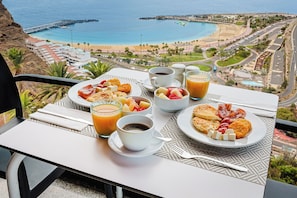 A table with breakfast items and drinks, overlooking a coastal view with buildings and a beach. - Hotel Chatur Palmera Mar (Mogan)