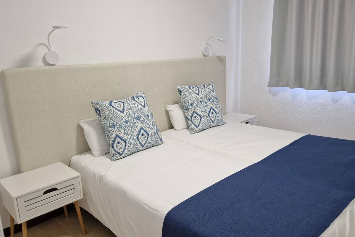 Una cama bien tendida con un cabecero blanco y azul estampada, una mesita de noche blanca con un cajón, y una manta azul.