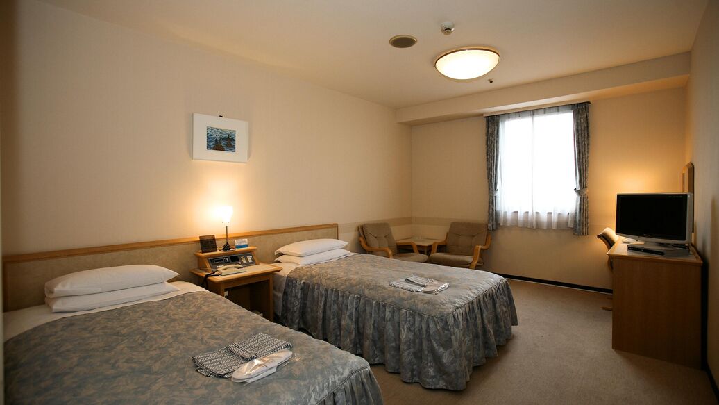A hotel room with two beds, a desk with a lamp, a chair, a television, and a window with curtains.
