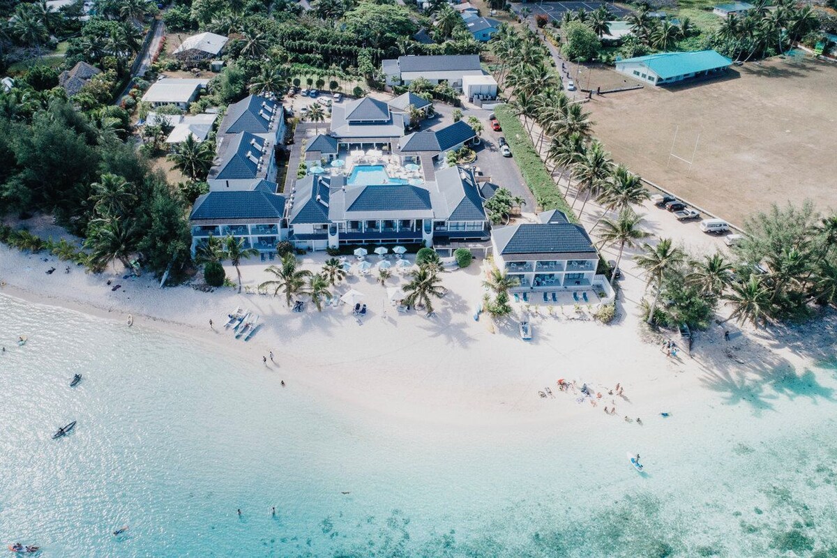 Vista aérea de un complejo turístico frente a la playa, con varias construcciones, una piscina y una playa de arena.