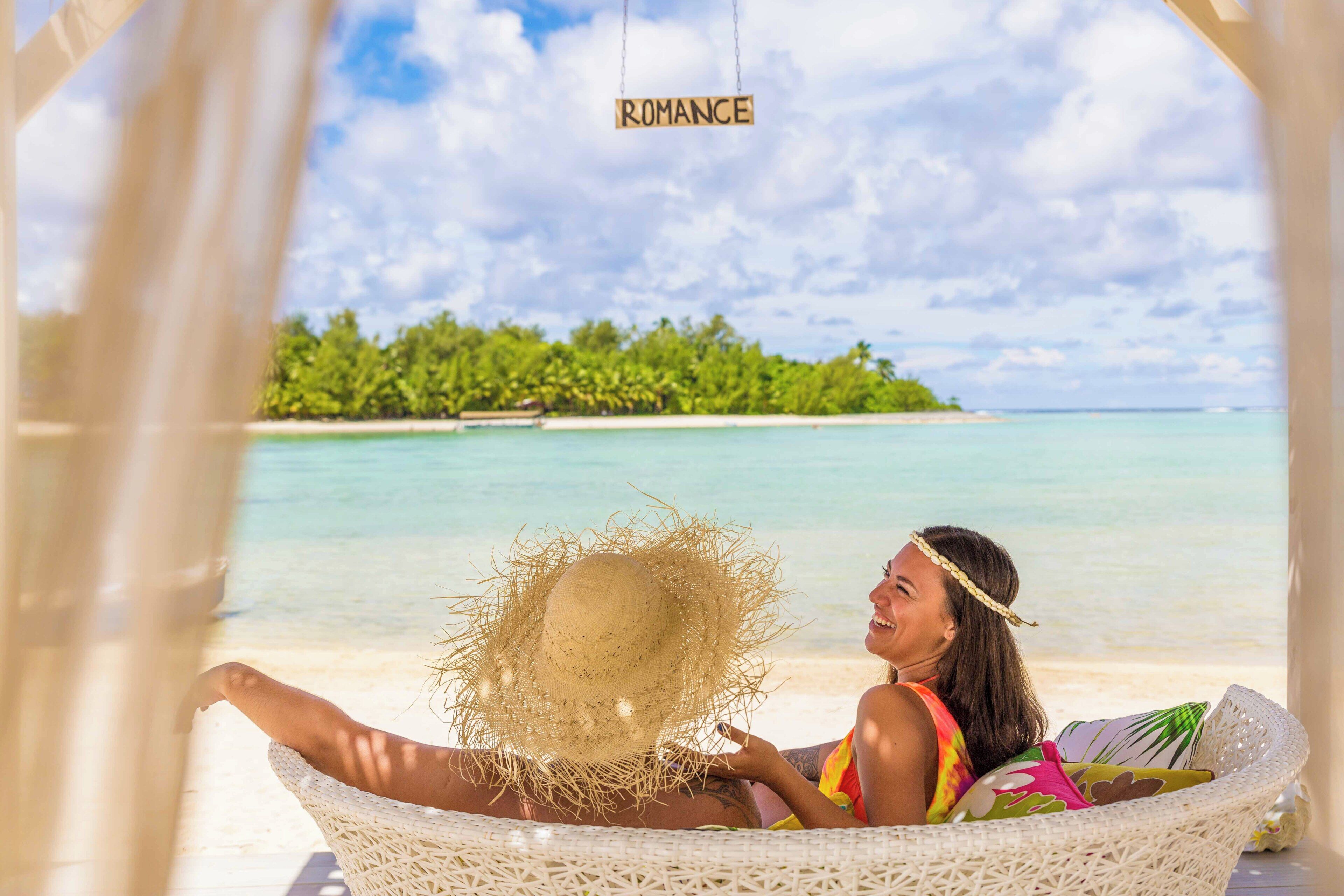 Una persona relajándose en una silla de mimbre con sombrero de paja, con vista a una playa, una pequeña isla y agua azul cristalina.