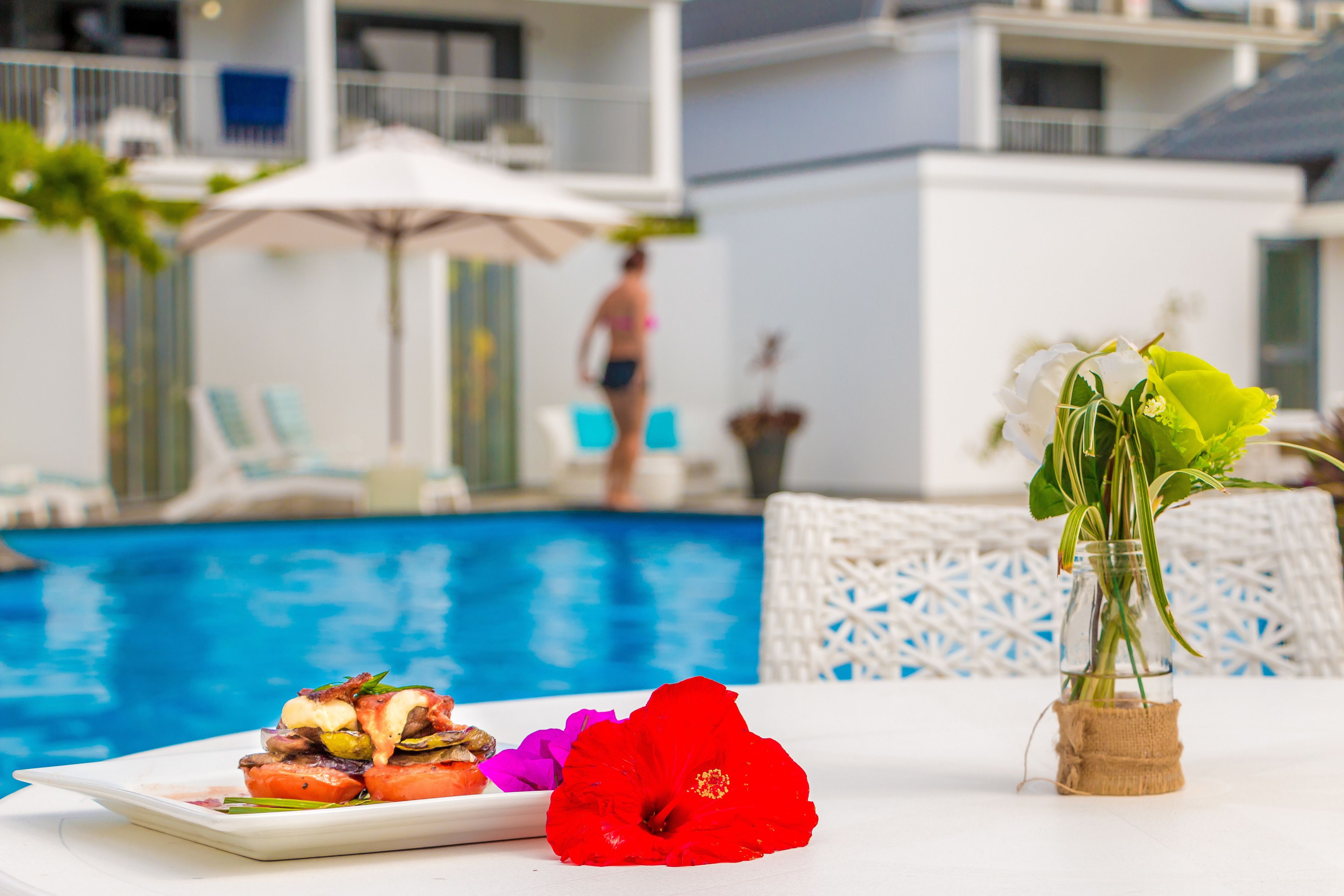 Une table placée au bord de la piscine, avec une assiette de nourriture, une fleur rouge et un vase contenant des plantes vertes.
