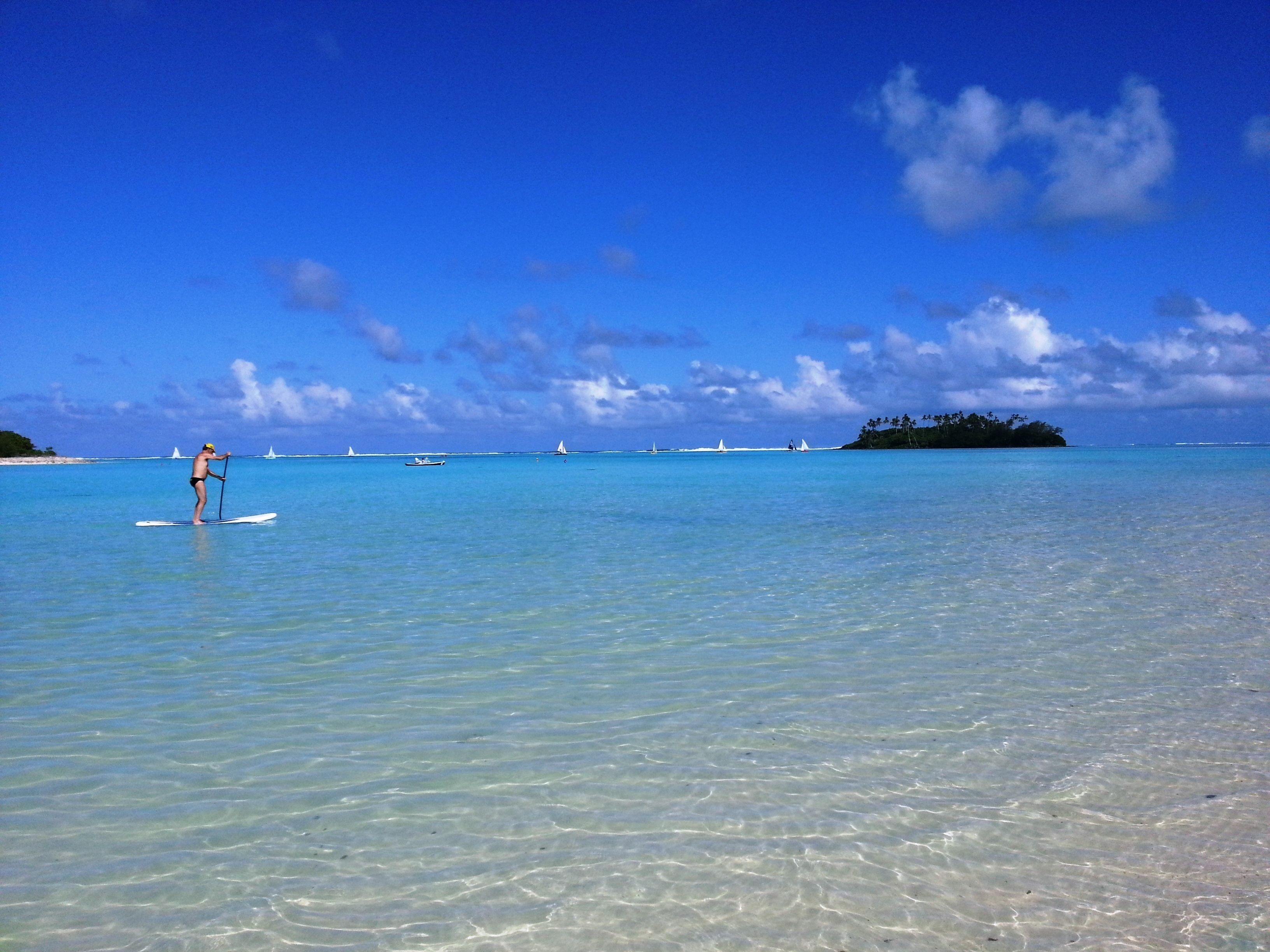 Ein unberührter tropischer Strand mit einer Person, die auf einem Stand-Up-Paddleboard fährt, ruhigem Wasser und fernen Segelbooten.