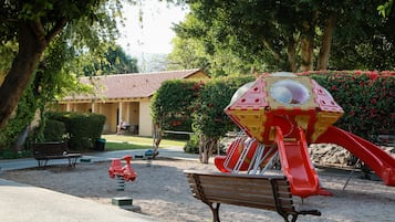 Enjoy a serene park setting with a play area featuring a slide and swing set, a bench for rest, and lush greenery.