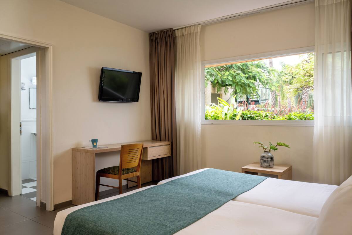 A hotel room with a bed, a desk with a chair, a television, a window with a view of greenery, and a small table with a vase.