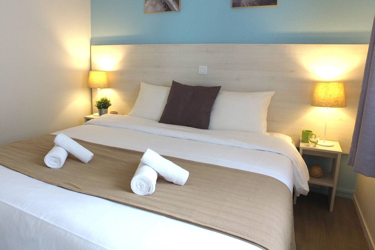 A neatly made bed with white linens, two rolled towels, and a wooden headboard. There are two bedside lamps and a small table with a green mug and a glass.