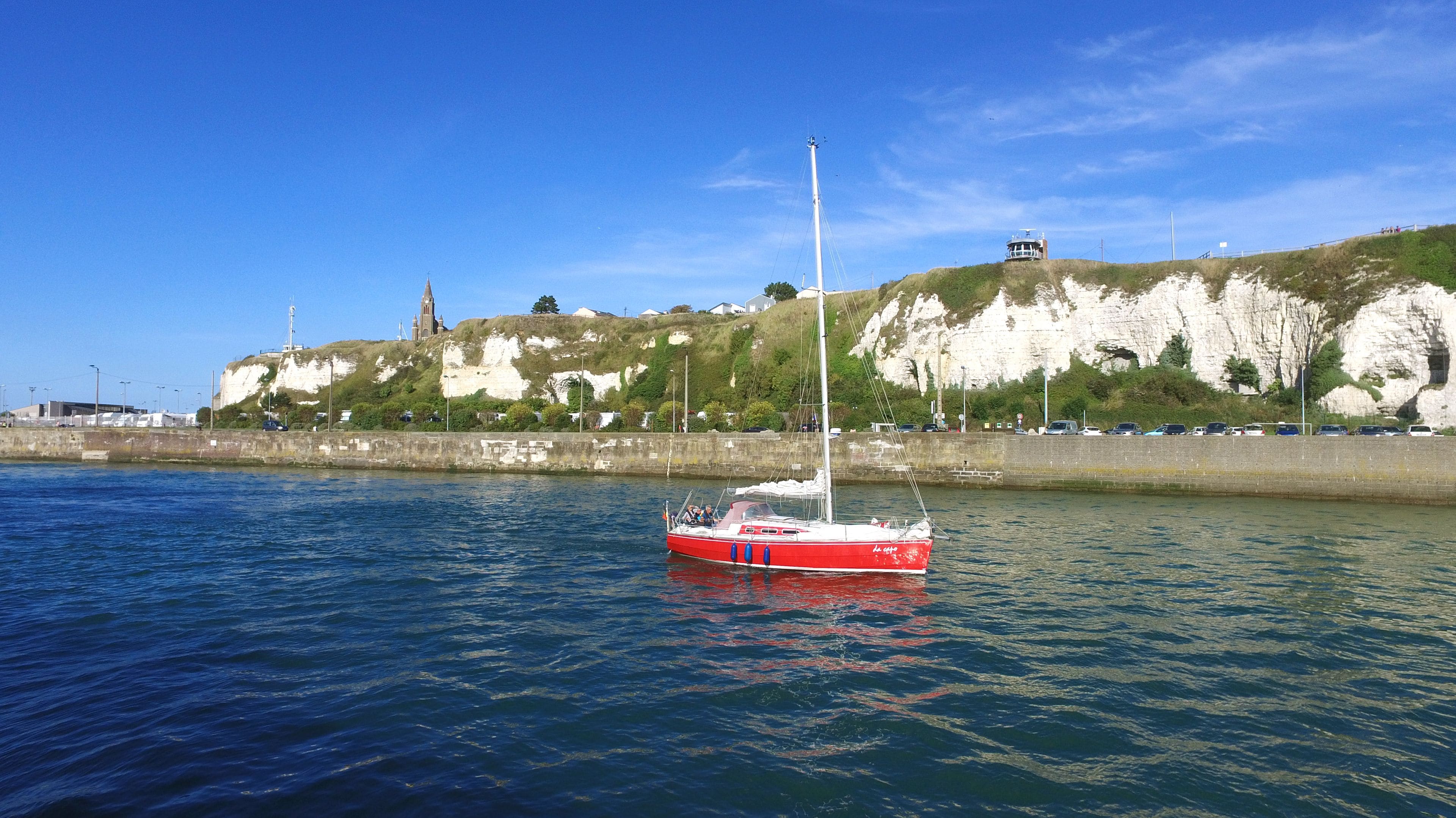 A red and white sailboat on calm water near a coastal area with cliffs and a lighthouse.
