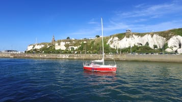 A red and white sailboat on calm water near a coastal area with cliffs and a lighthouse.