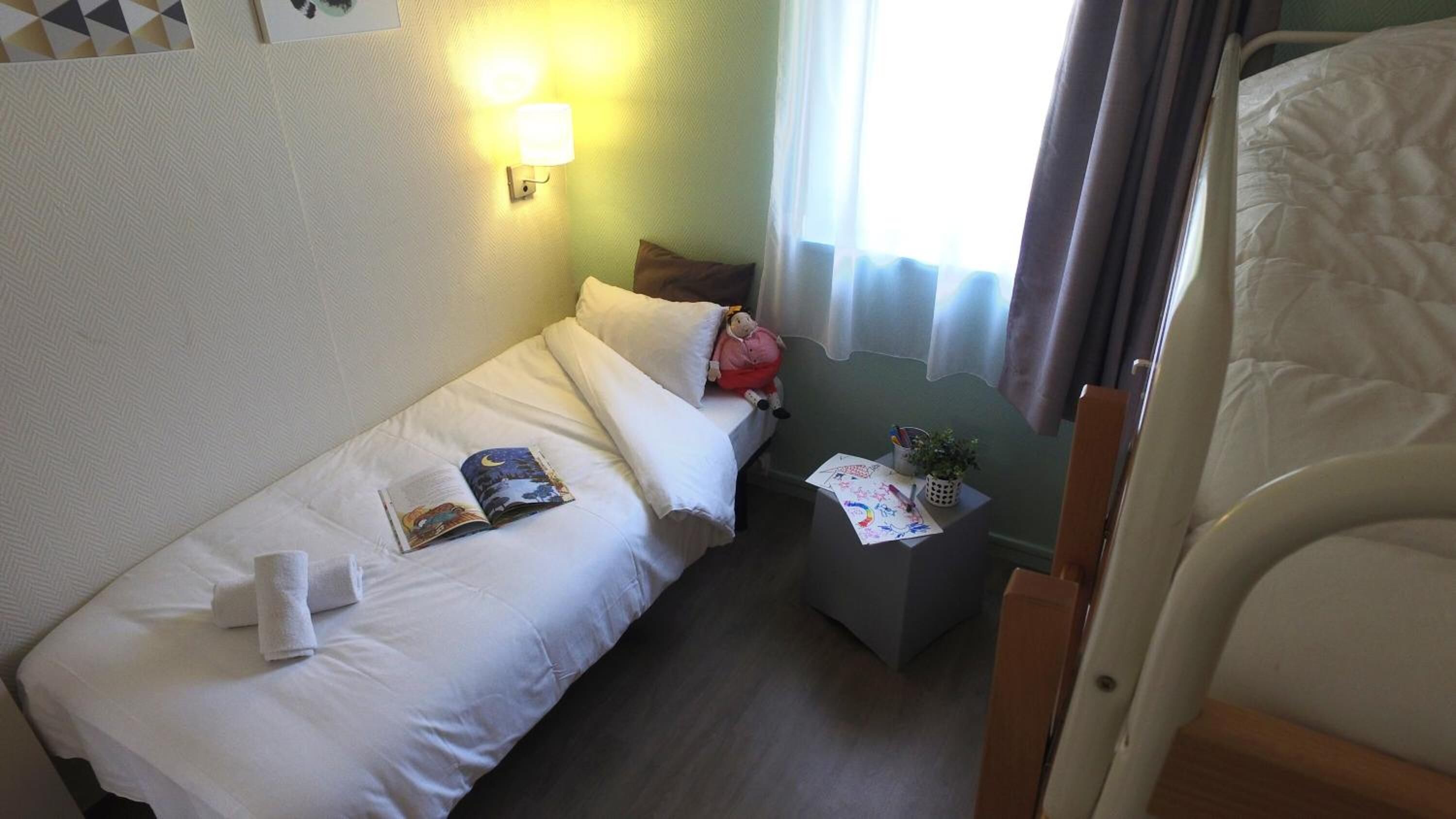 A small, neatly made bed with a pillow and a book, a bedside table with a lamp, and a window with curtains.