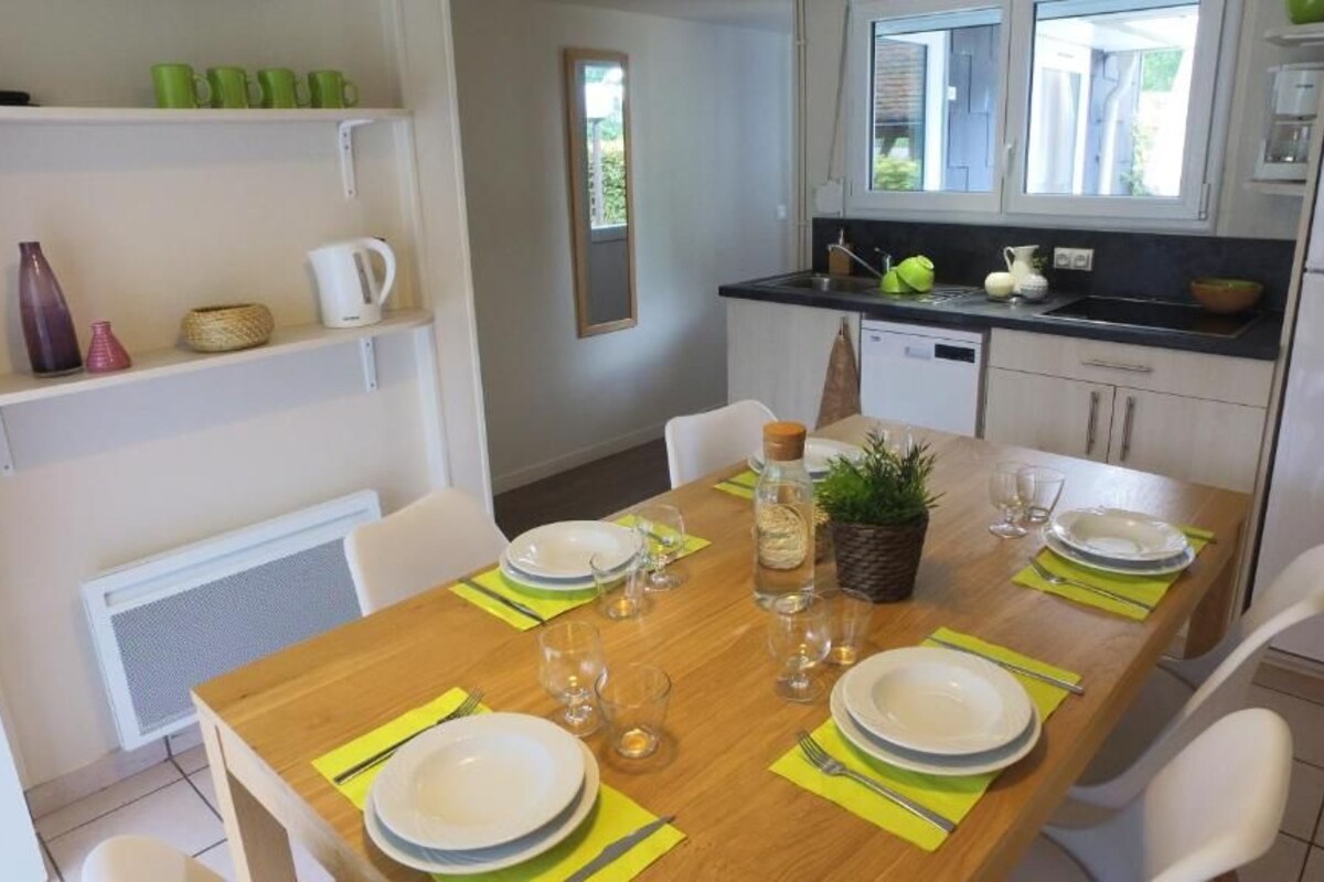 Un comedor con una mesa de madera dispuesta para cuatro personas, sillas blancas y una cocina al fondo.