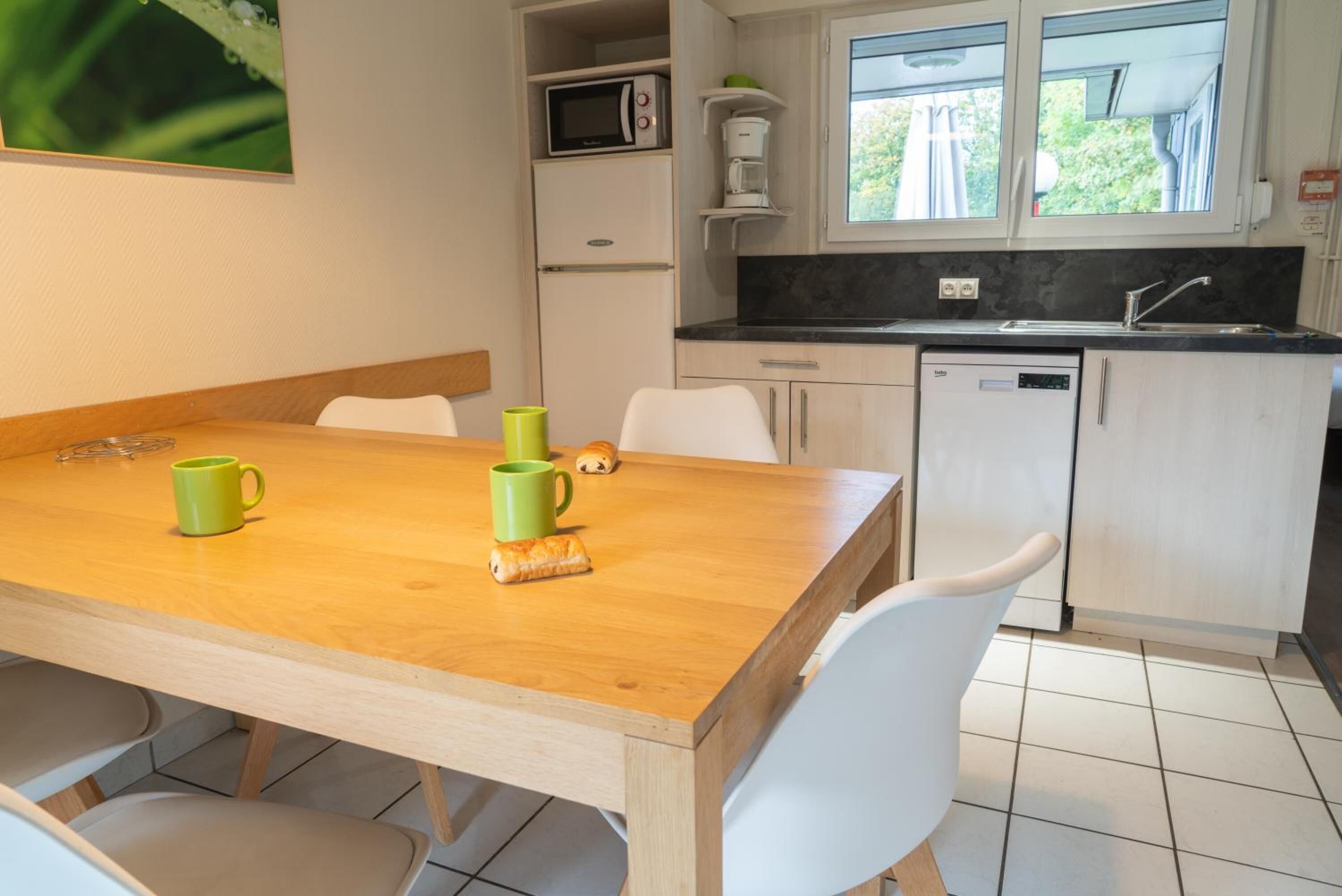 A compact kitchen with a wooden table, white chairs, a microwave, a coffee maker, and a dishwasher.