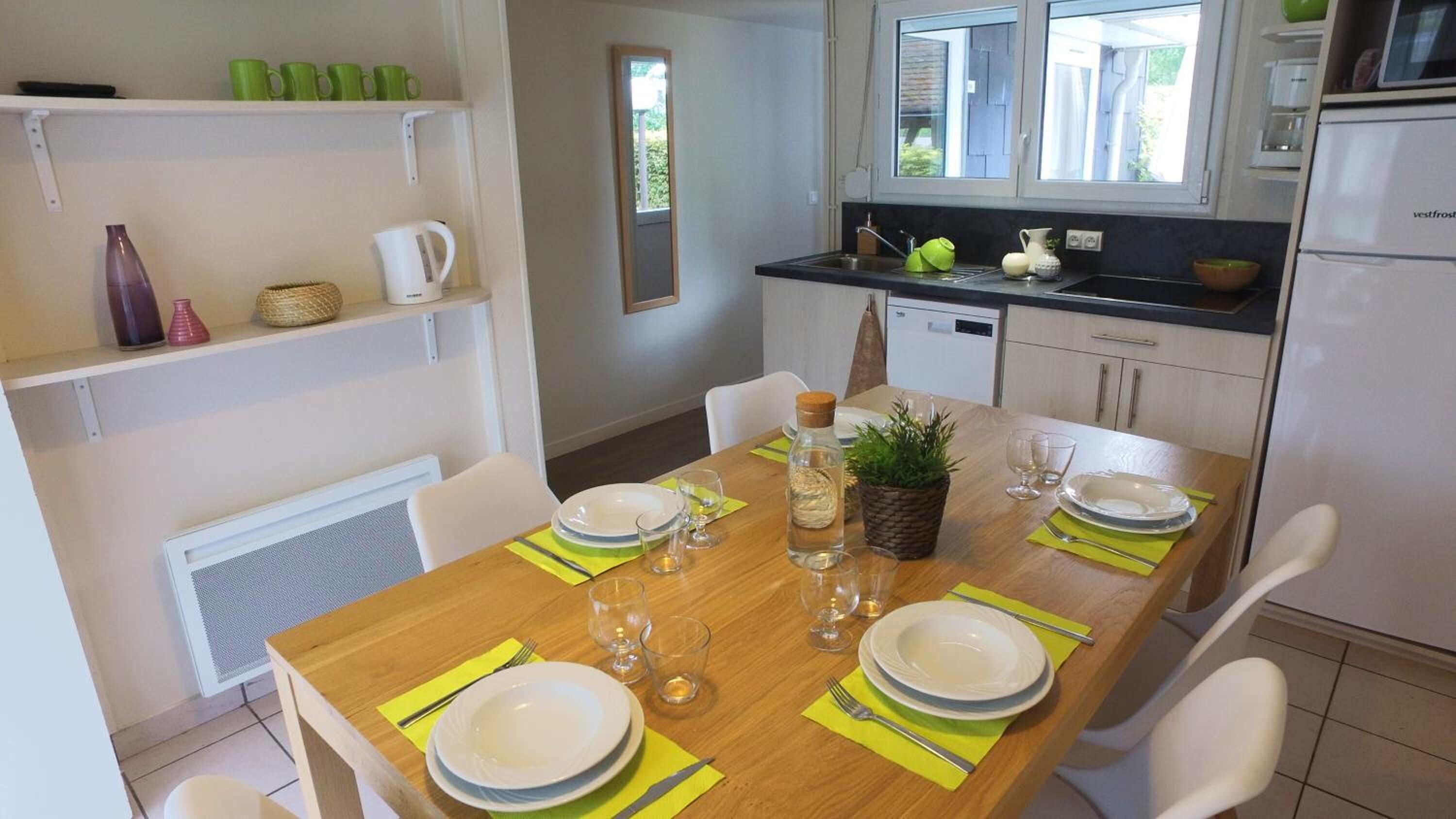 Uma sala de jantar com mesa de madeira posta para quatro pessoas, cercada por cadeiras brancas. Há uma área de cozinha com geladeira, pia e armários.