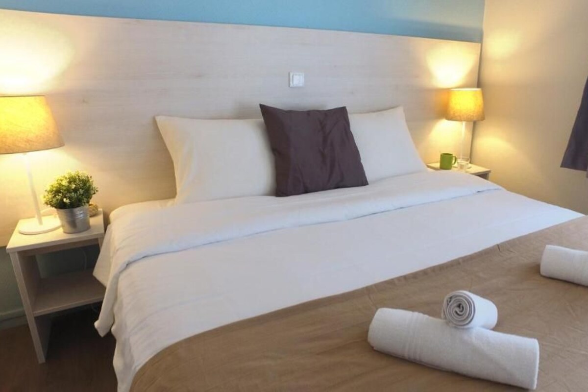 Habitación de hotel con cama, dos lámparas de noche, una mesita de noche con una planta y una ventana con cortinas.
