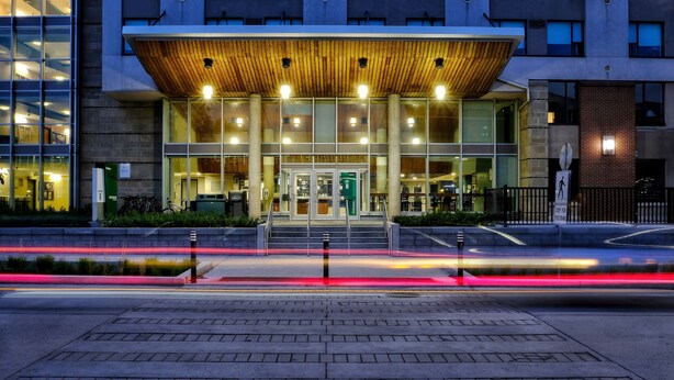 A modern building with a wooden canopy entrance, large glass windows, and a visible sign indicating a parking area. - Residence & Conference Centre - Ottawa West (Ottawa)