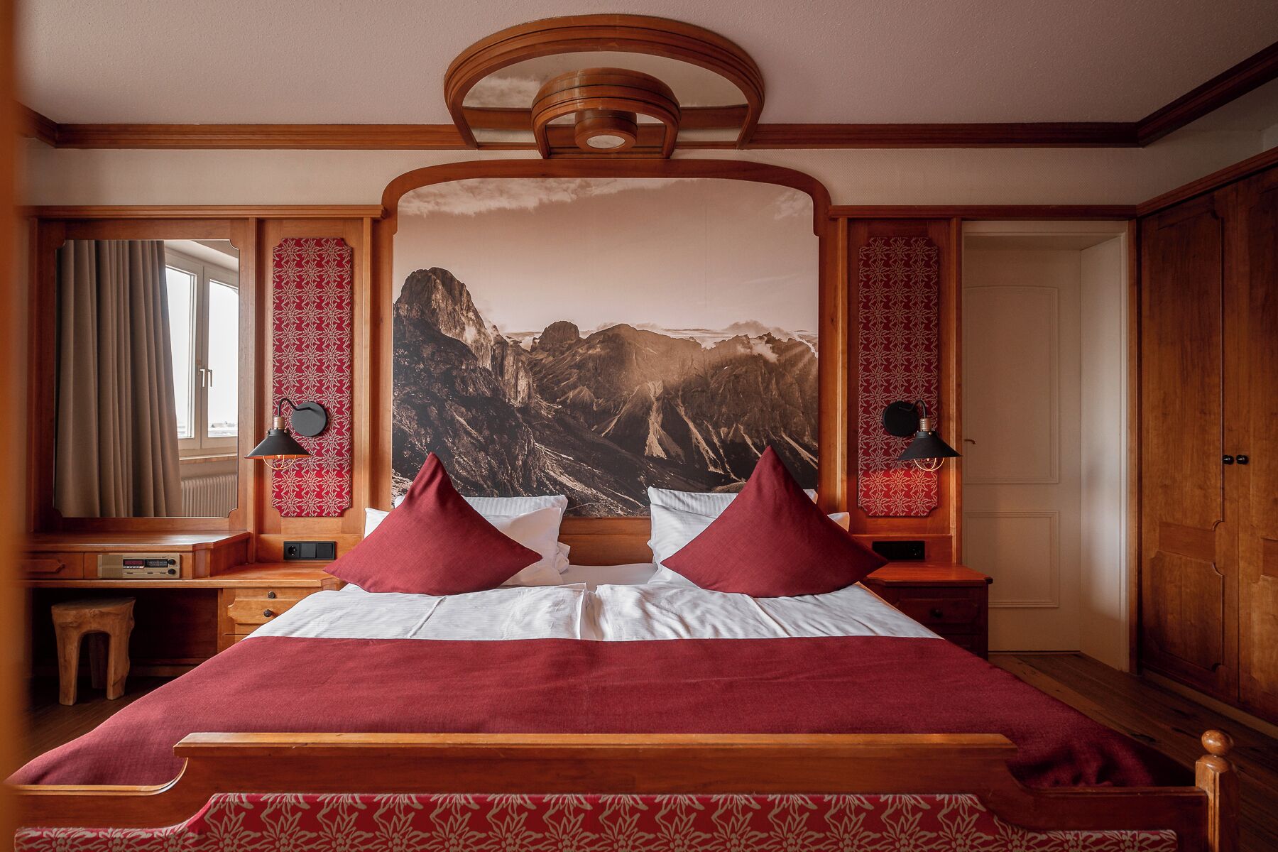Une chambre d’hôtel avec un grand lit, deux lampes de chevet, une tête de lit en bois, une grande fenêtre avec des rideaux et une fresque murale représentant un paysage de montagne.