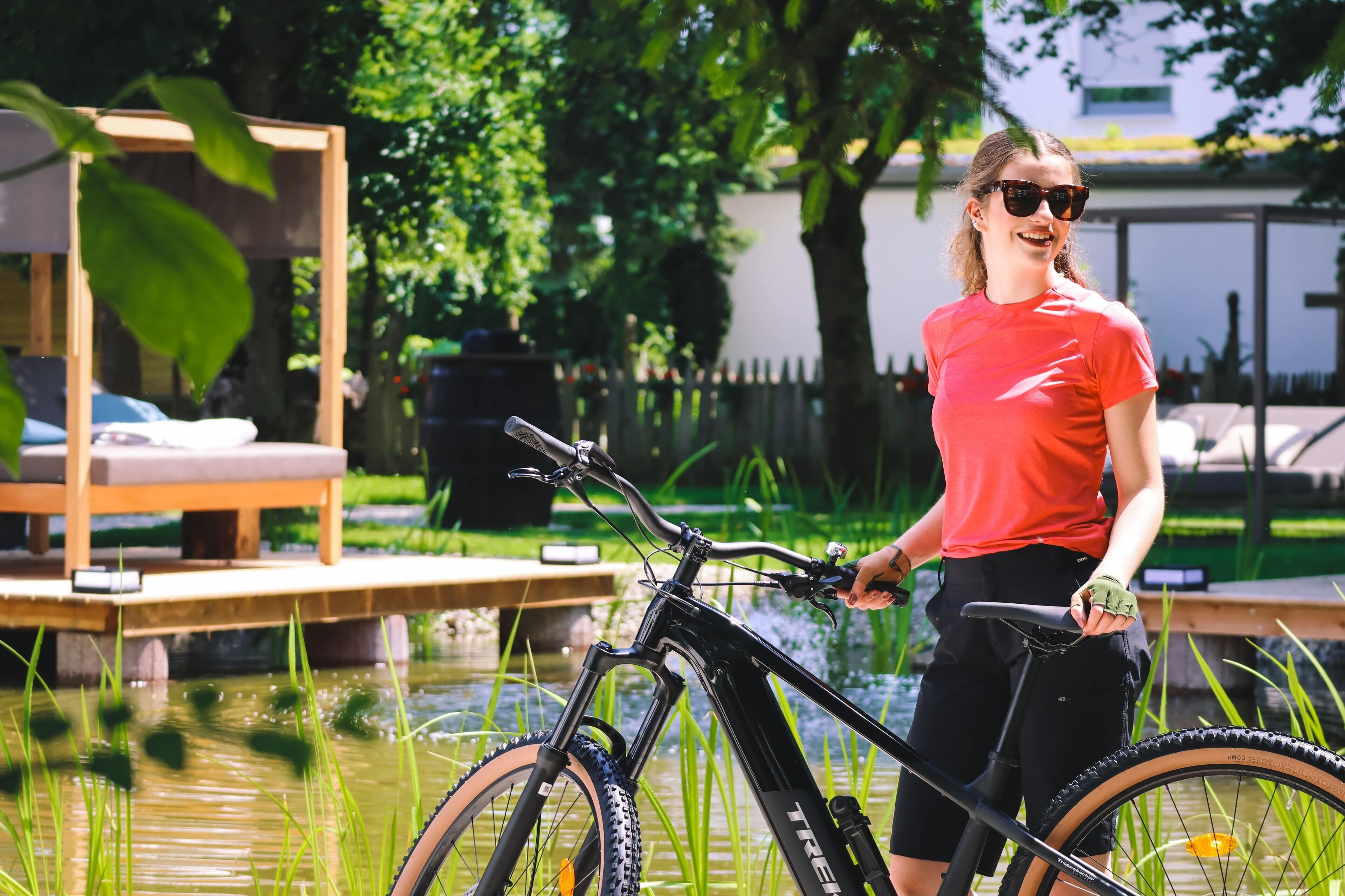 Uma pessoa ao lado de uma bicicleta em área de jardim com deck de madeira e lago.