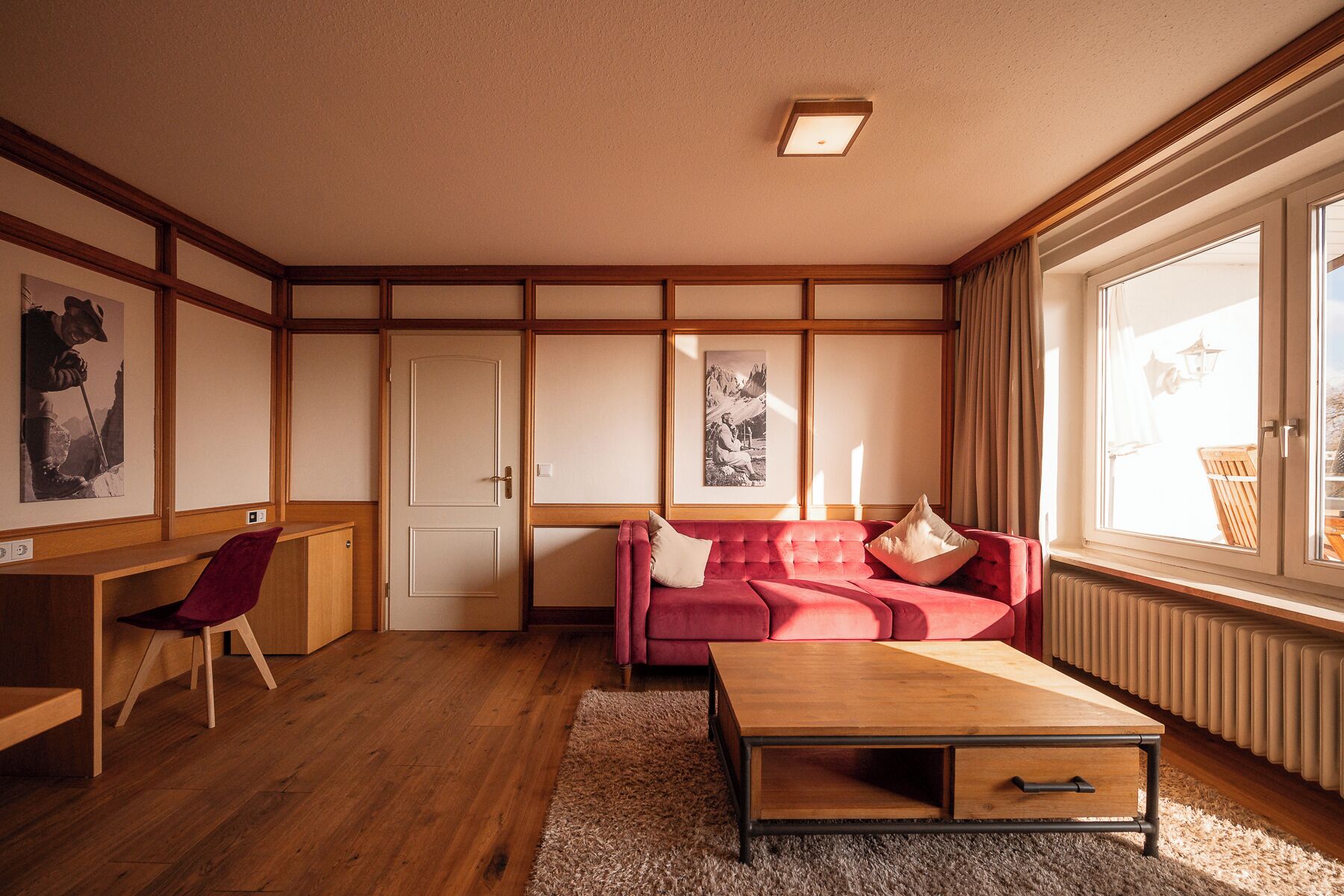 A room with a wooden floor, a red sofa, a wooden coffee table, a desk, and framed pictures on the wall.