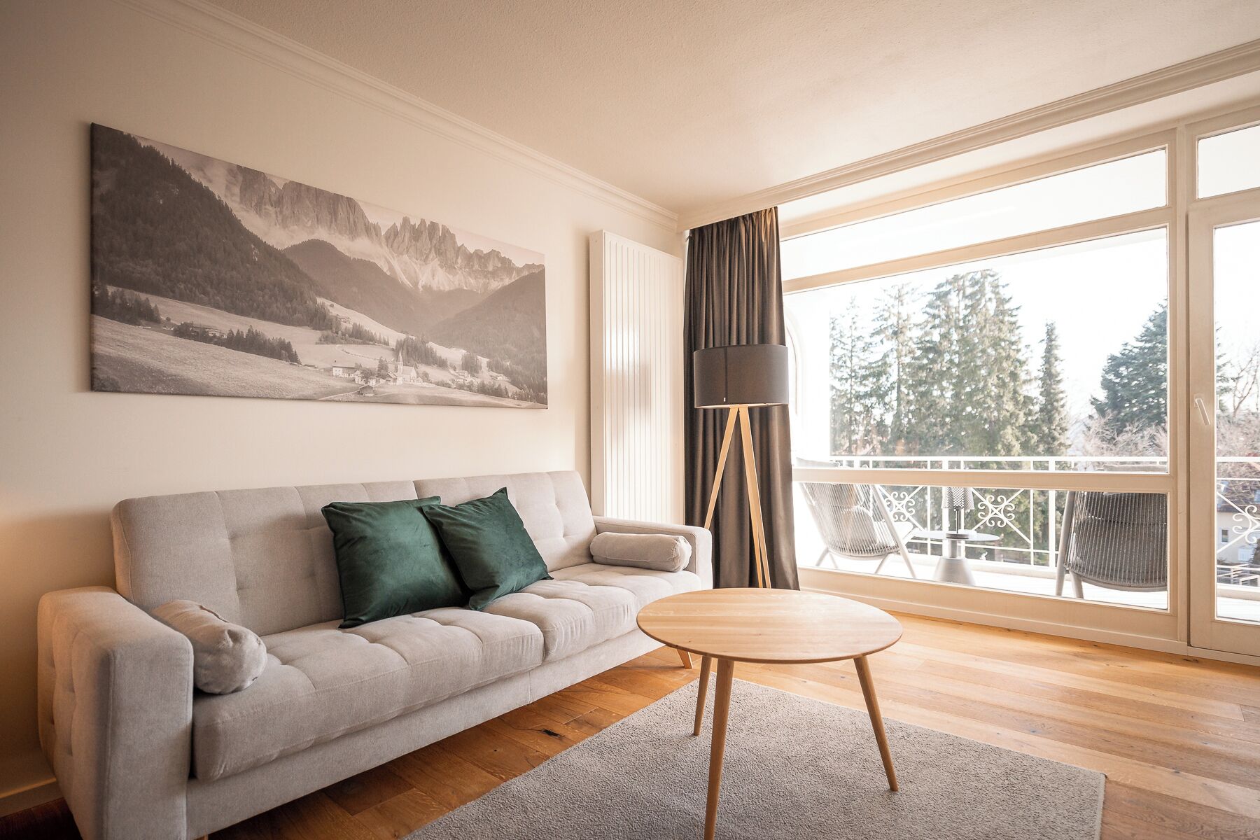 A modern living room with a sofa, a round wooden table, a floor lamp, and a large window with a view of trees.
