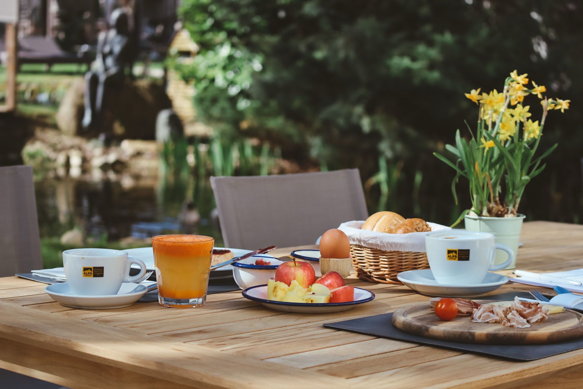 Une table de petit-déjeuner avec une tasse de café, un verre de jus d’orange, une assiette de fruits, un panier de pain et une planche à découper avec de la viande.
