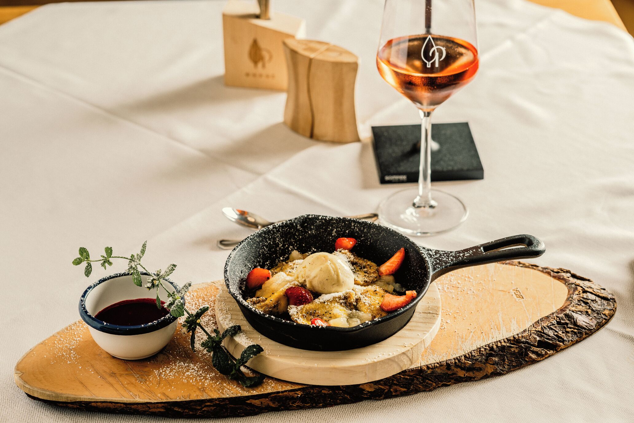 Dessert in einer Pfanne auf einer Holzplatte, dazu eine kleine Schale Soße und ein Glas Wein auf einem Tisch.