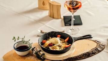 Dessert in einer Pfanne auf einer Holzplatte, dazu eine kleine Schale Soße und ein Glas Wein auf einem Tisch.