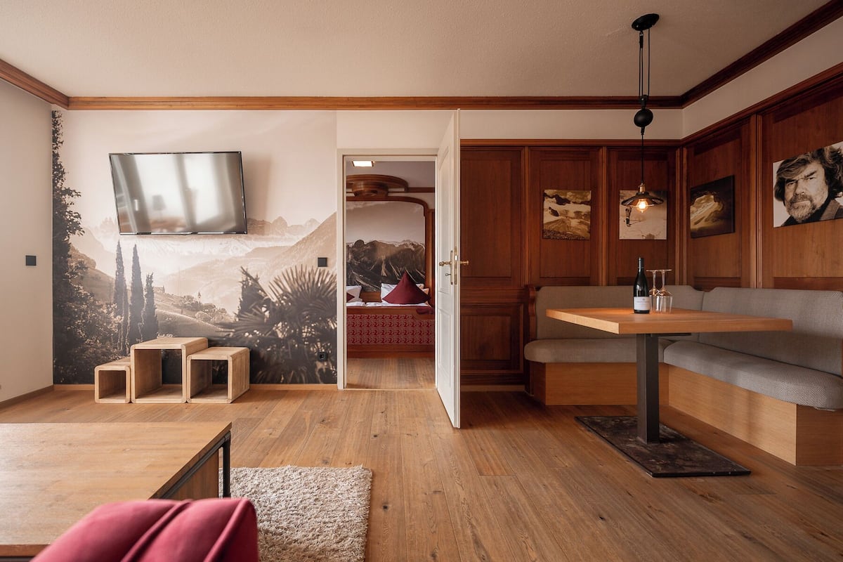 Una habitación con piso de madera, un sofá, una mesa con una botella y vasos, un televisor montado en la pared y una puerta que da a otra habitación.