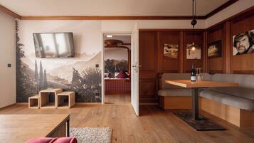 Una habitación con piso de madera, un sofá, una mesa con una botella y vasos, un televisor montado en la pared y una puerta que da a otra habitación.