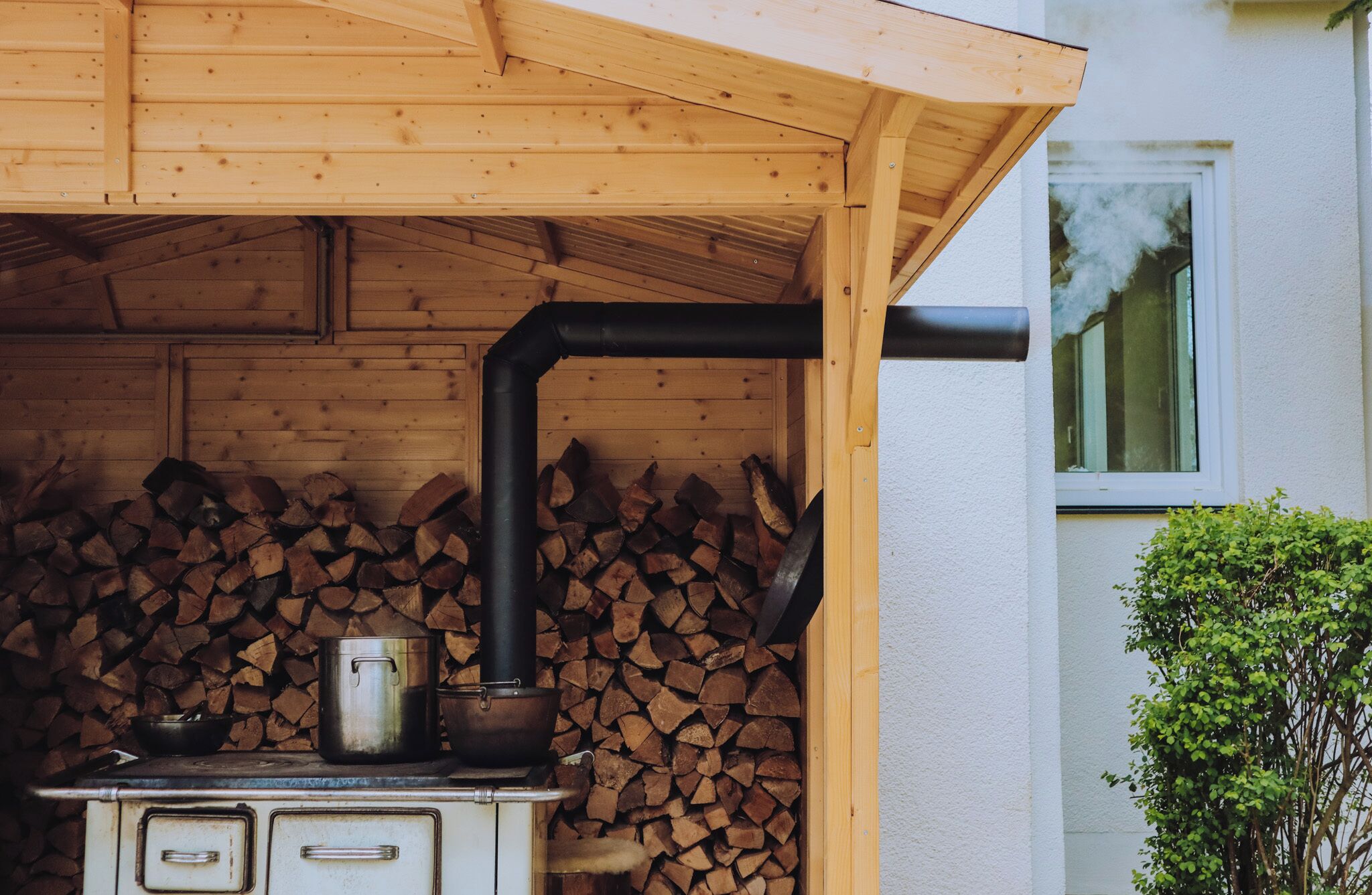 Ein Holzofen mit Schornstein, ein Topf darauf und ein Holzstoß an einer Holzwand.